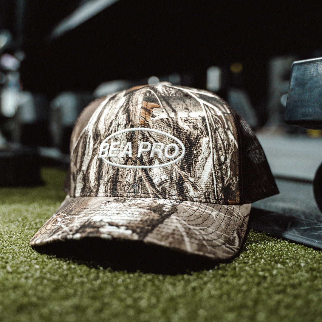 GBRS Group Be A Pro Trucker Hat in Realtree Camo - pictured on the floor of a gym