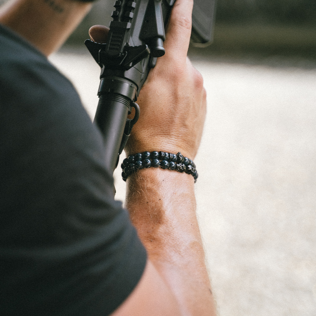 A close-up lifestyle shot of a man's arm wearing a black beaded bracelet while holding a black rifle. The background is a soft-focus outdoor setting.