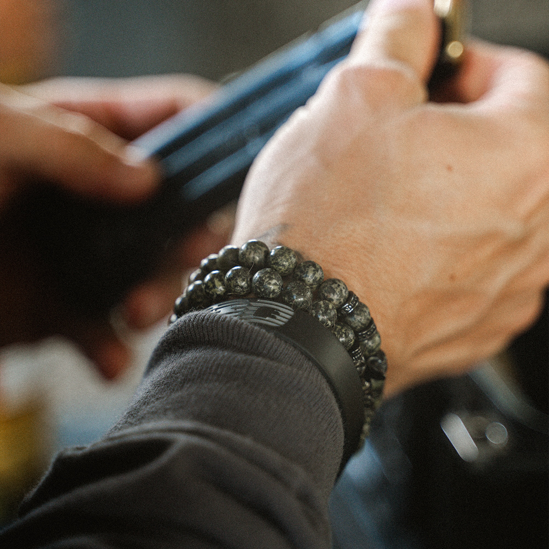 A close-up of a person's wrist wearing a black beaded bracelet with metallic silver-gray beads and a black leather wristband, while holding a dark object.