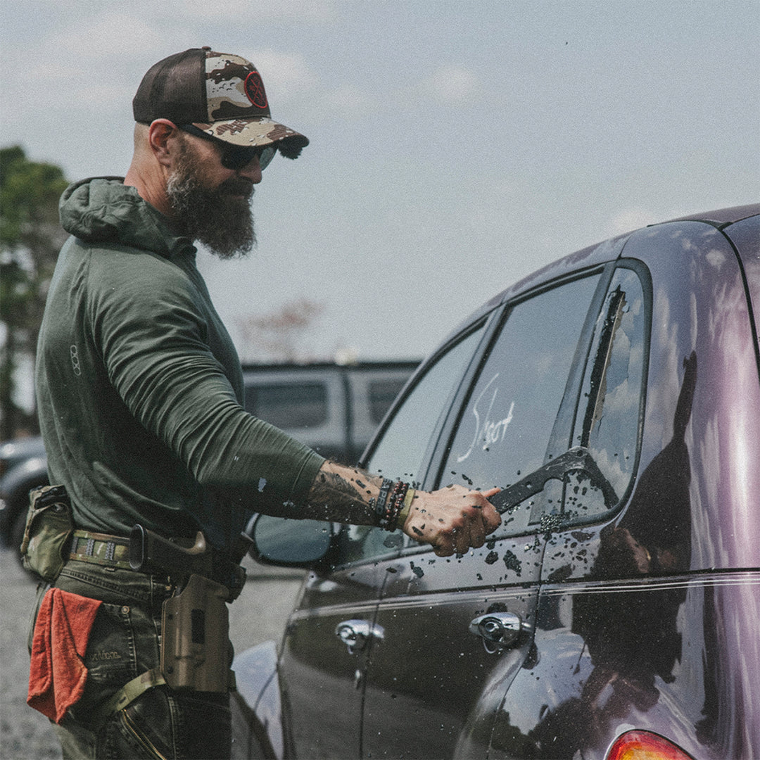 Man with a beard and camouflage hat, breaking a car window with a sharp, black metal tool bar.