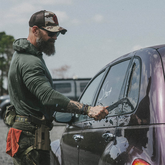 Man with a beard and camouflage hat, breaking a car window with a sharp, black metal tool bar.