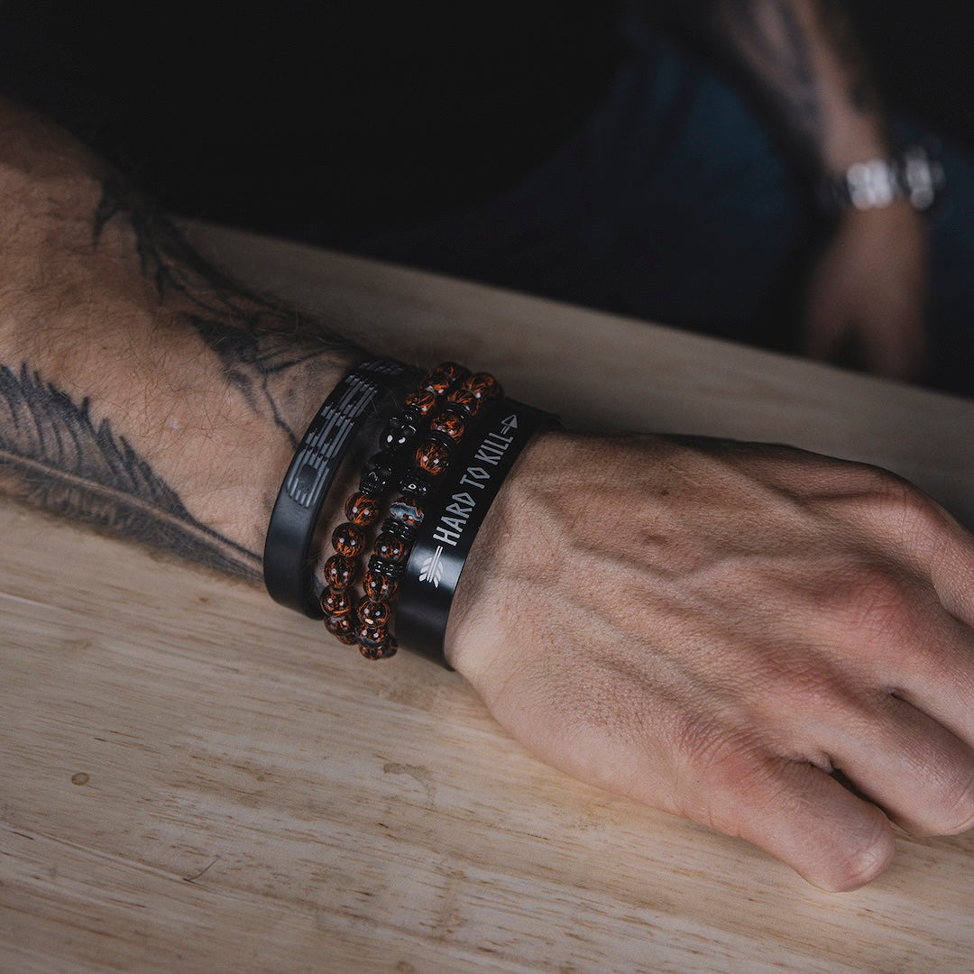 Man wearing GBRS Group Hard to Kill Cuff on wrist among other bracelets and accessories with his arm resting on a table