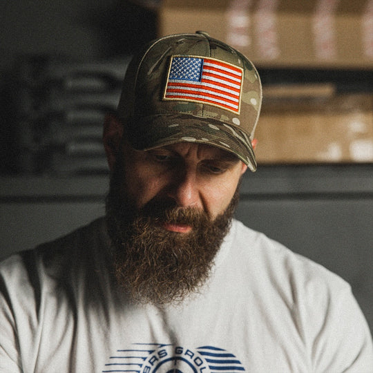 A bearded man wearing a Multicam camouflage trucker hat with an American flag in the center and a white t-shirt, standing in a dimly lit, rustic indoor setting.