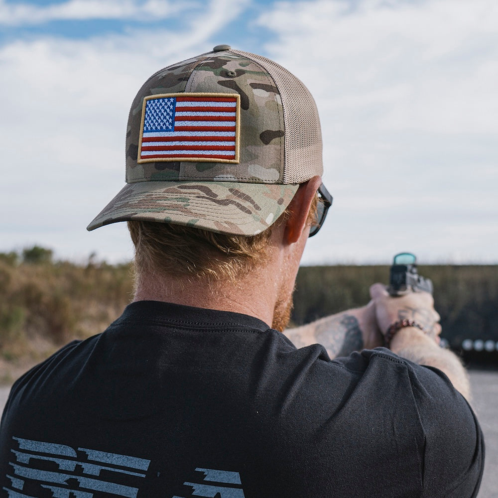 Man wearing a Multicam camo trucker hat featuring an American flag, with a pistol at an outdoor shooting range.