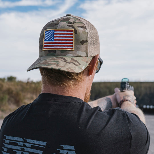 Man wearing a Multicam camo trucker hat featuring an American flag, with a pistol at an outdoor shooting range.