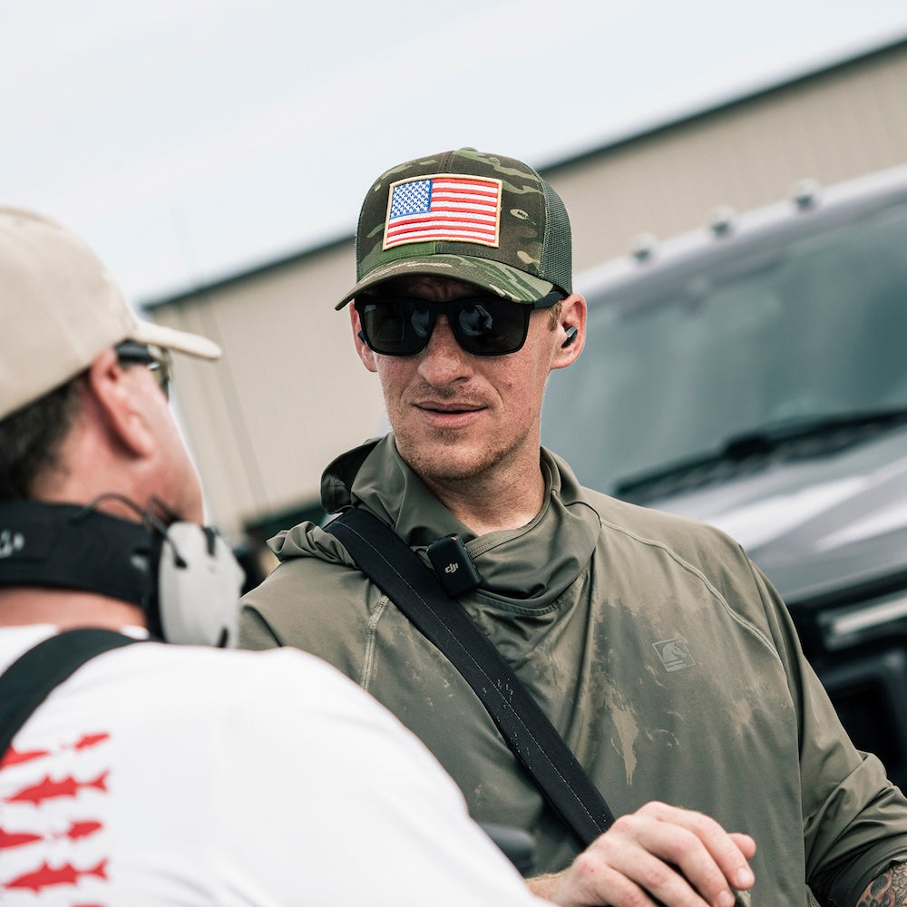 A man wearing a camouflage trucker hat with an American flag patch, black sunglasses, and an olive green hooded shirt is shown in conversation outdoors near a vehicle.