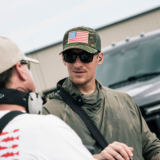 A man wearing a camouflage trucker hat with an American flag patch, black sunglasses, and an olive green hooded shirt is shown in conversation outdoors near a vehicle.