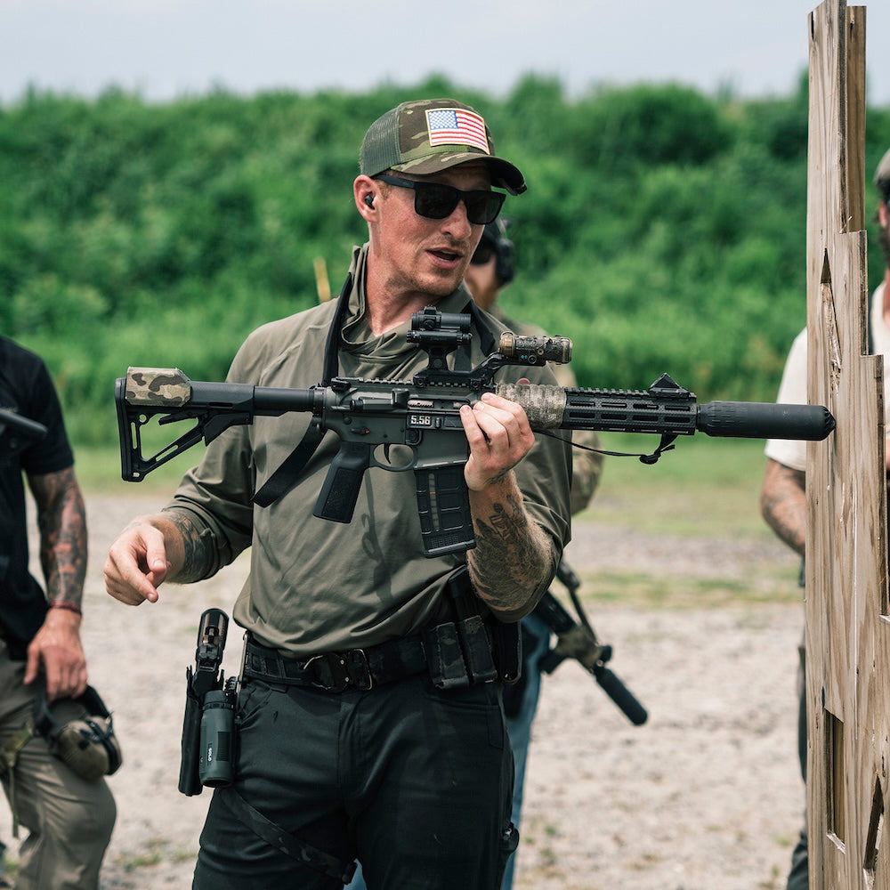 A man wearing a camouflage hat with an American flag patch, sunglasses, and an olive green shirt stands at an outdoor shooting range holding a black tactical rifle with a mounted scope and suppressor. Other people and a wooden target stand are visible in the background.