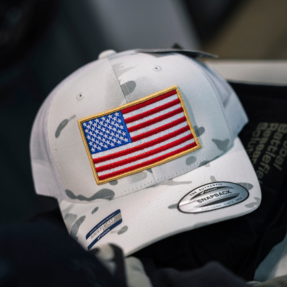 A white Multicam camouflage trucker hat featuring an embroidered American flag patch on the front, with a visible Snapback brand sticker on the brim.