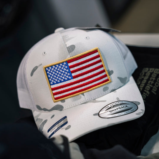 A white Multicam camouflage trucker hat featuring an embroidered American flag patch on the front, with a visible Snapback brand sticker on the brim.