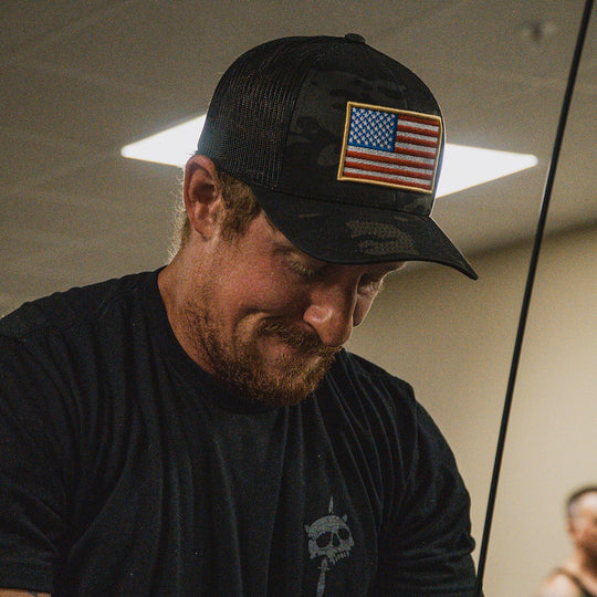 A man wearing a black camouflage trucker hat with a US flag patch on the front, paired with a black t-shirt, captured in an indoor setting.