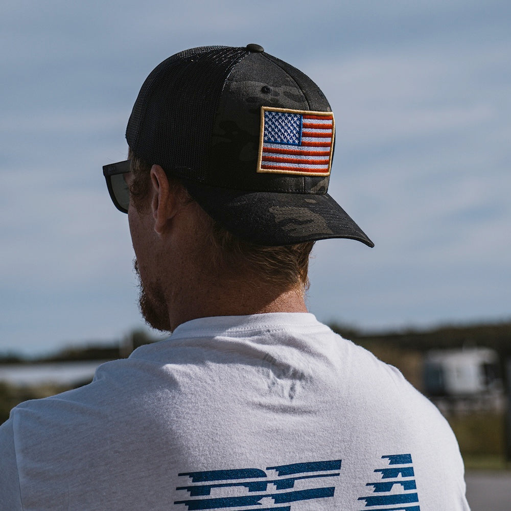 A man viewed from behind wearing a black camouflage trucker cap with an embroidered American flag patch on the side, paired with a white t-shirt.