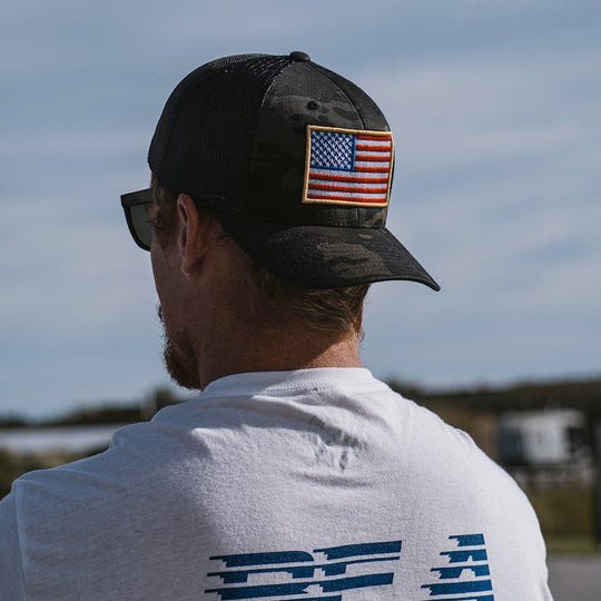 A man viewed from behind wearing a black camouflage trucker cap with an embroidered American flag patch on the side, paired with a white t-shirt.