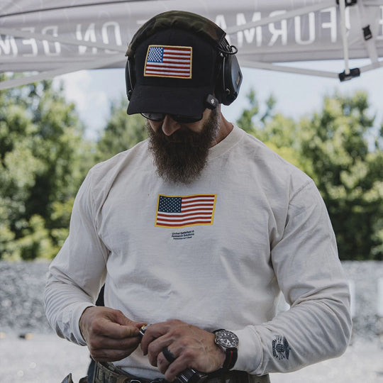 Man wearing the GBRS Group Patriotism Trucker Hat, featuring an American flag patch, and a long-sleeve shirt with a similar patch, outdoors.