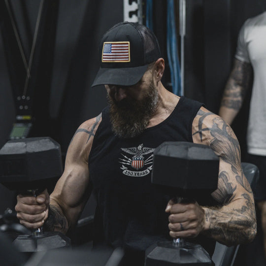Man with tattoos lifting dumbbells in a gym setting, while wearing the GBRS Group Patriotism Trucker Hat.