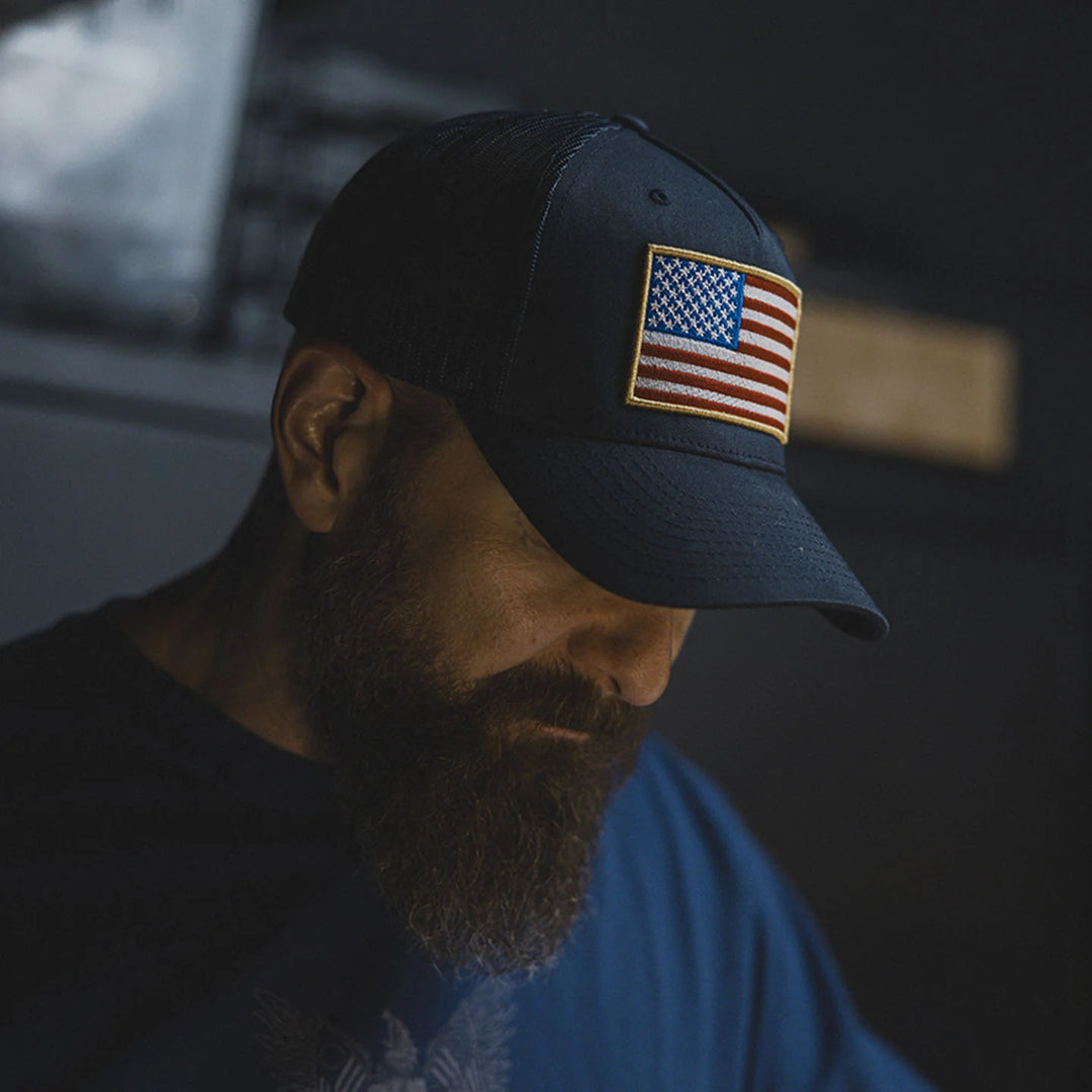 Man wearing a navy blue cap with an American flag patch, the GBRS Group Patriotism Trucker Hat, in a workshop setting.