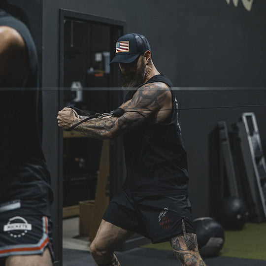 Man with tattoos performing an exercise in a gym setting, while wearing the GBRS Group Patriotism Trucker Hat.