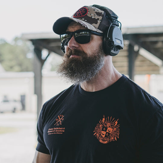 Man wearing a black t-shirt with a GBRS Group design, sunglasses, and a hat outdoors.