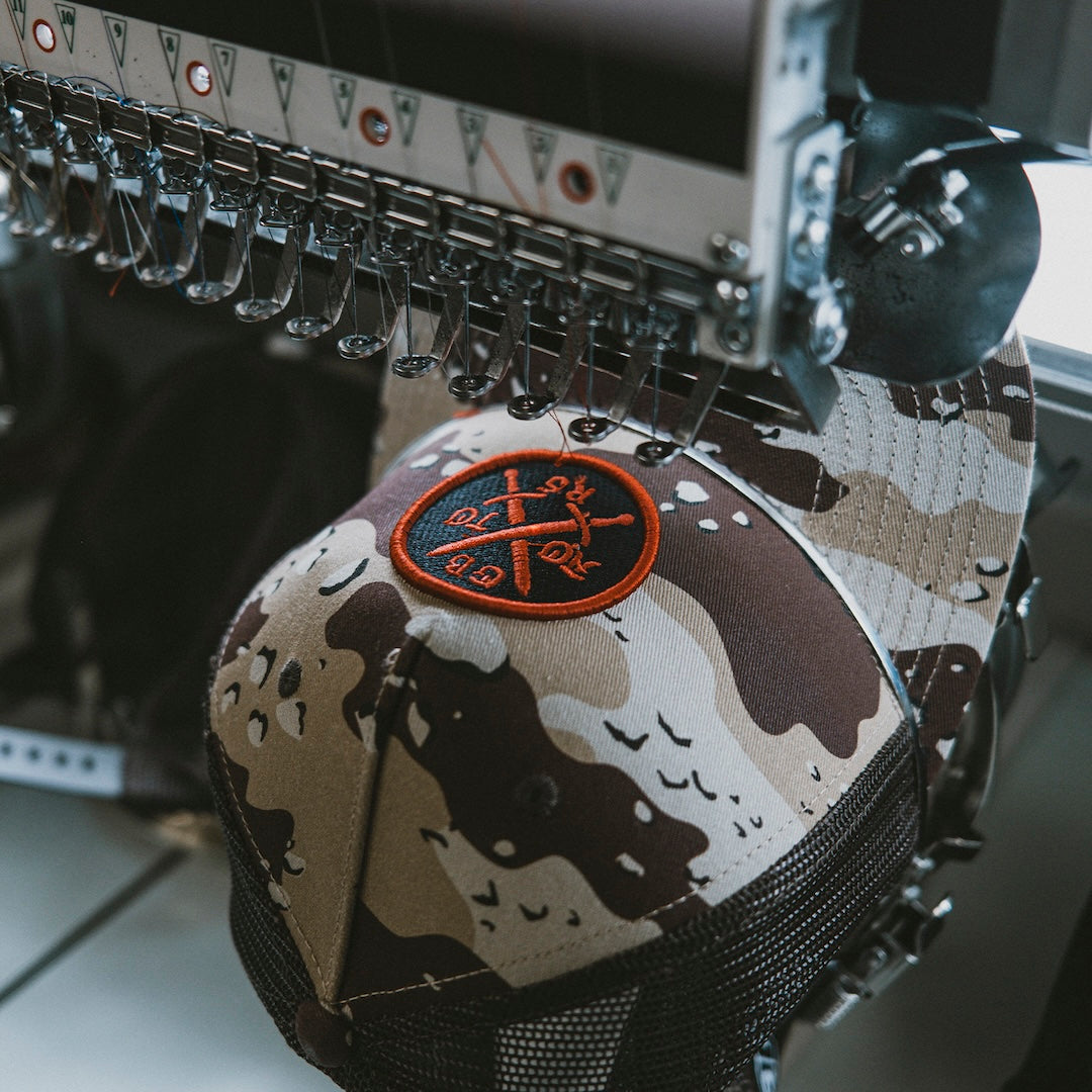 Camouflage-patterned hat with embroidered patch design in the middle getting worked on and made on an embroidery machine.