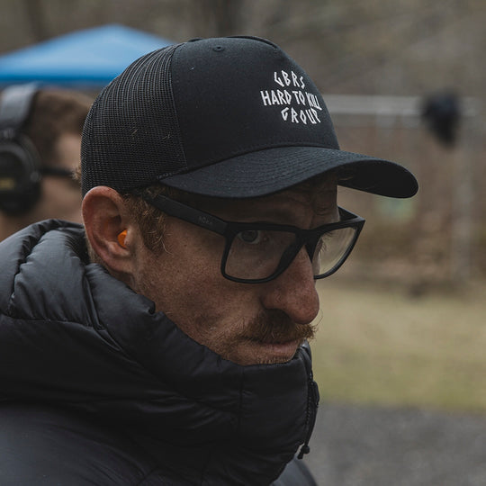 Man wearing GBRS Hard to Kill Trucker Hat with clear eyeglasses in an outdoor setting
