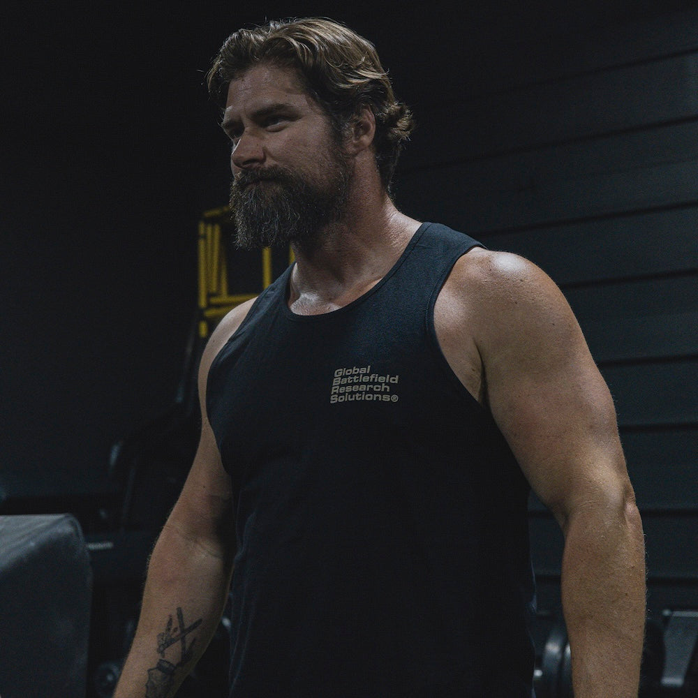 A muscular bearded man in a gym wearing a black tank top with the text 'Global Battlefield Research Solutions' printed in white on the chest.