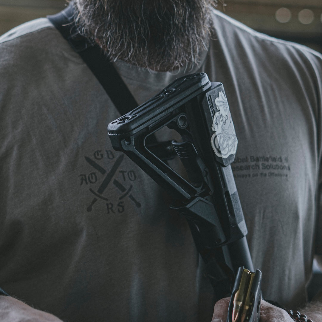 Man holding a rifle on a sling, wearing a gray t-shirt with text and designs.