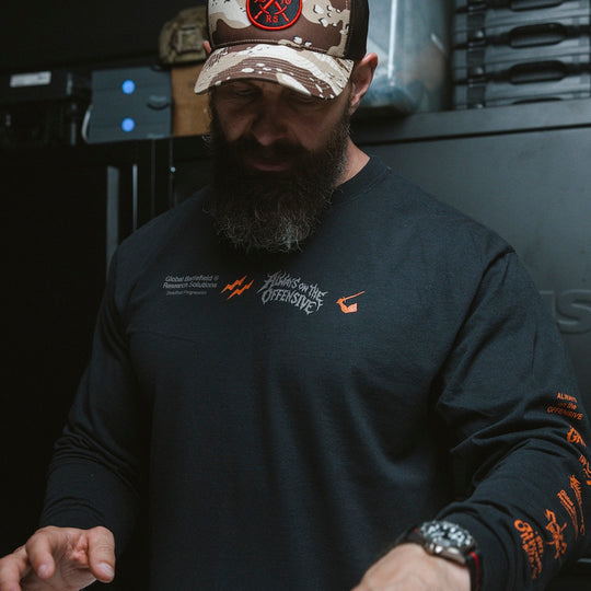 Man wearing a black long-sleeve shirt with logos and text, standing in an indoor setting.