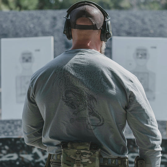 Man wearing a gray long sleeve, ear protection, and a hat, standing in front of target practice boards.