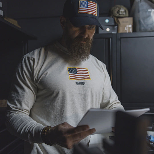 Man wearing a cap and GBRS Group Front and Center Long Sleeve Shirt with American flag patches, holding a tablet.