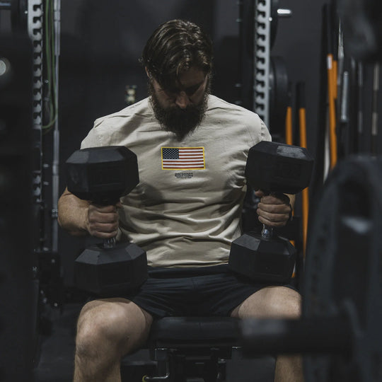 Man lifting dumbbells in a gym setting wearing the GBRS Group Front and Center Cream Short Sleeve Shirt, with an American flag in the center of the shirt.