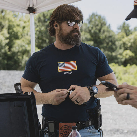 Man wearing the GBRS Group Front and Center Navy Short Sleeve Shirt with an American flag in the center, at an outdoor range.
