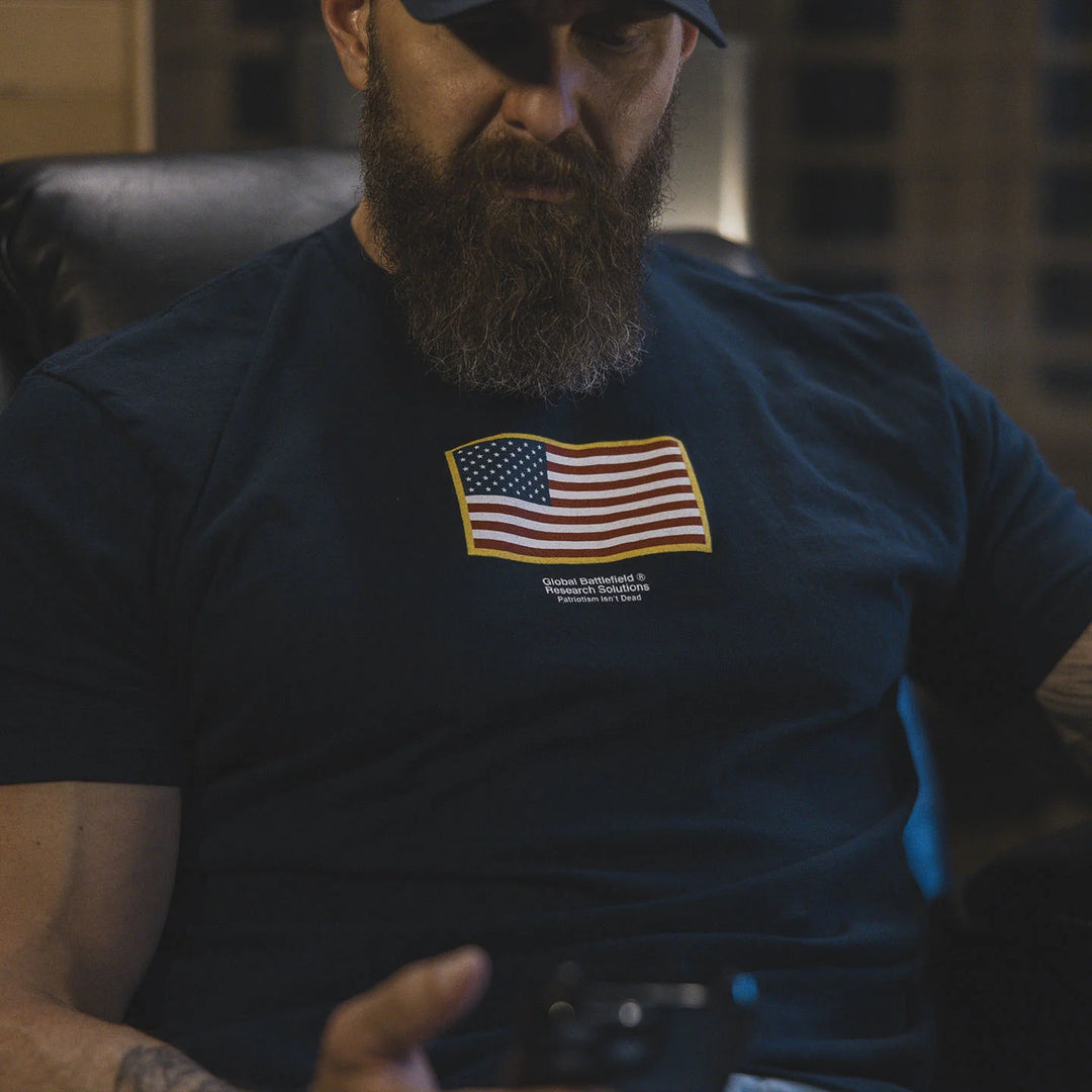 Man wearing the GBRS Group Front and Center Navy Short Sleeve Shirt with an American flag design, sitting in a dimly lit room.