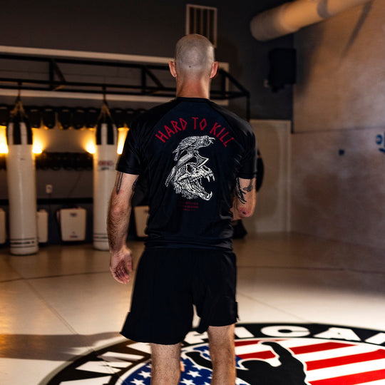 Image of a man wearing the GBRS Group Hard to Kill Rash Guard in a gym setting showing the design on the back