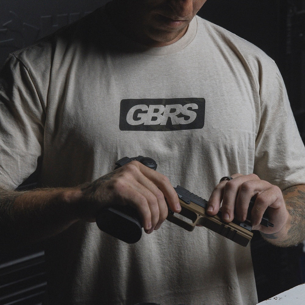 A man wearing a light beige t-shirt with a black and white 'GBRS' logo on the chest, holding a handgun in a tactical room.