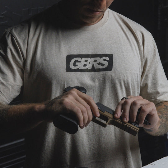 A man wearing a light beige t-shirt with a black and white 'GBRS' logo on the chest, holding a handgun in a tactical room.