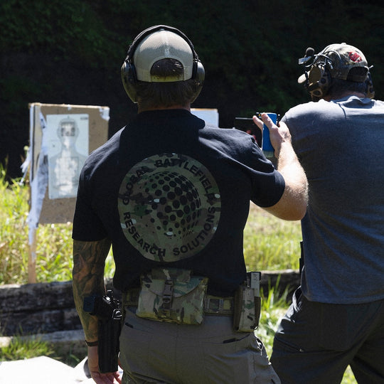 Man viewed from behind at an outdoor range, wearing the GBRS Group Infiltration Short Sleeve Shirt. The graphic design on the back is visible and features a large multicam-patterned design.