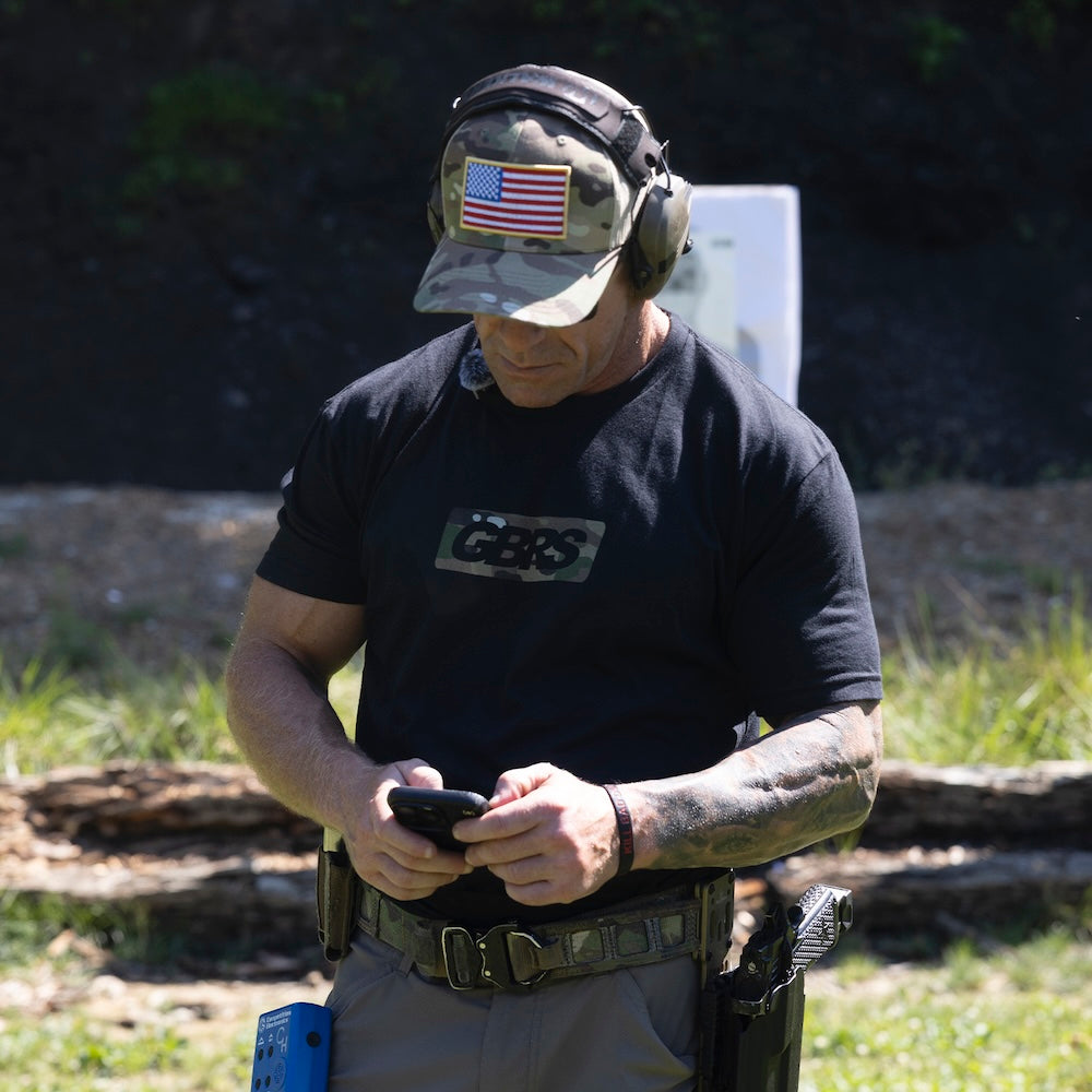 A man wearing a black GBS t-shirt, a camouflage cap with an American flag patch, and ear protection stands at an outdoor shooting range looking at a phone.