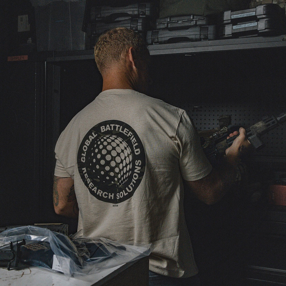 Person wearing the cream-colored GBRS Group Infiltration Short Sleeve Shirt while holding a tactical rifle, standing in a store room full of tactical equipment. 
