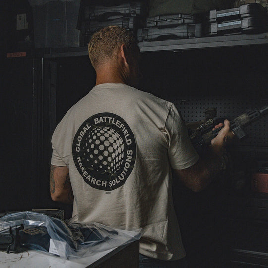 Person wearing the cream-colored GBRS Group Infiltration Short Sleeve Shirt while holding a tactical rifle, standing in a store room full of tactical equipment. 