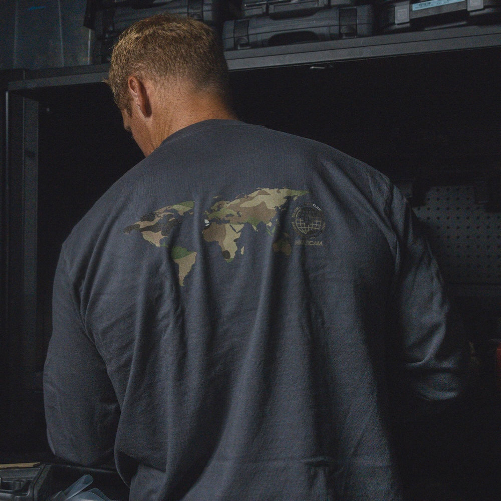 A man viewed from behind wearing the GBRS Group International Long Sleeve Shirt, featuring a camouflage world map graphic and a small globe logo on the back, standing in a storage room.