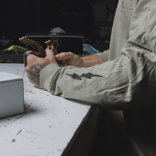 A close up image of a person holding an optic mount, showing the sleeve thunderbolt design of the GBRS Group International Long Sleeve Shirt.