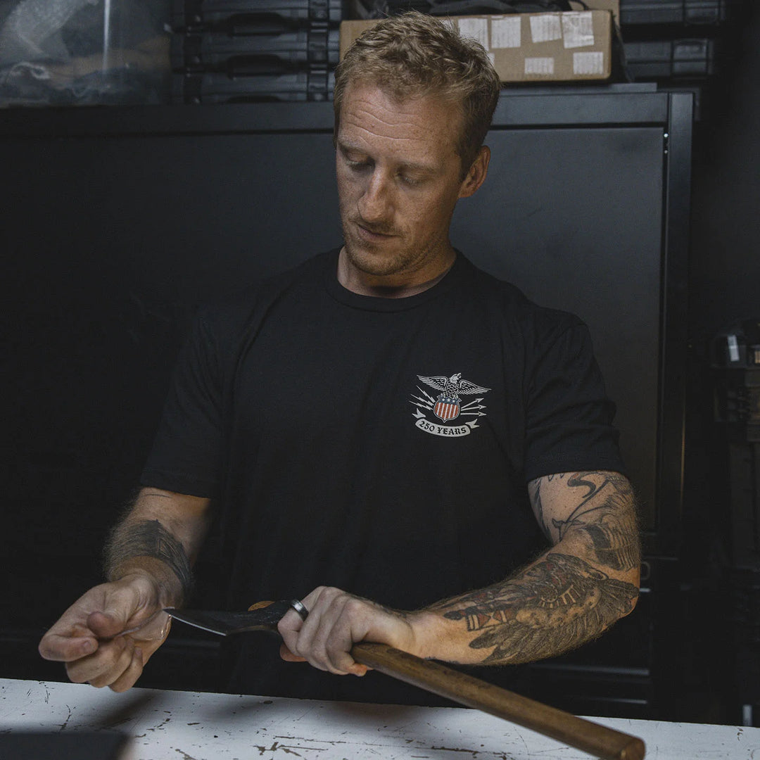 Man with tattoos holding a knife and wooden object in a workshop setting, wearing the GBRS Group Lead and Lace Short Sleeve Shirt.