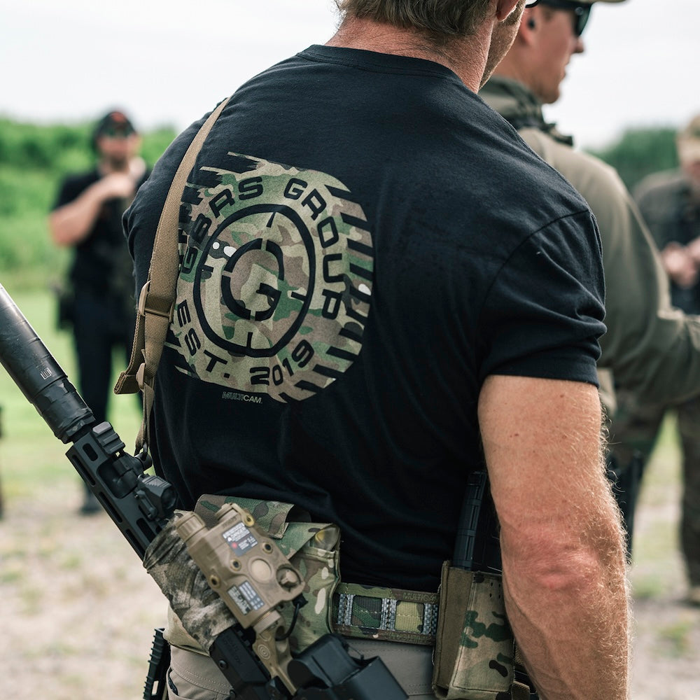 Person wearing the GBRS Group Obscured Short Sleeve Shirt, showing the design on the back. Person is standing at an outdoor shooting range with a tactical rifle slung around his back.