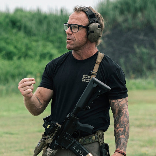 Man wearing a black t-shirt with a tactical rifle around his chest, while speaking outdooors.
