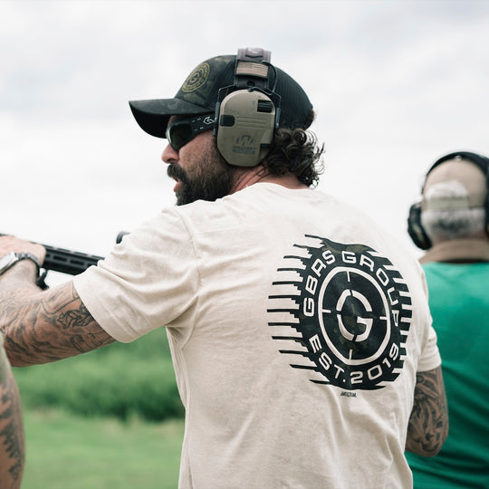 A man at an outdoor shooting range wearing a cream t-shirt with a large black circular 'GBRS Group' logo on the back, a black cap, and ear protection. Another person is visible in the background on the right.