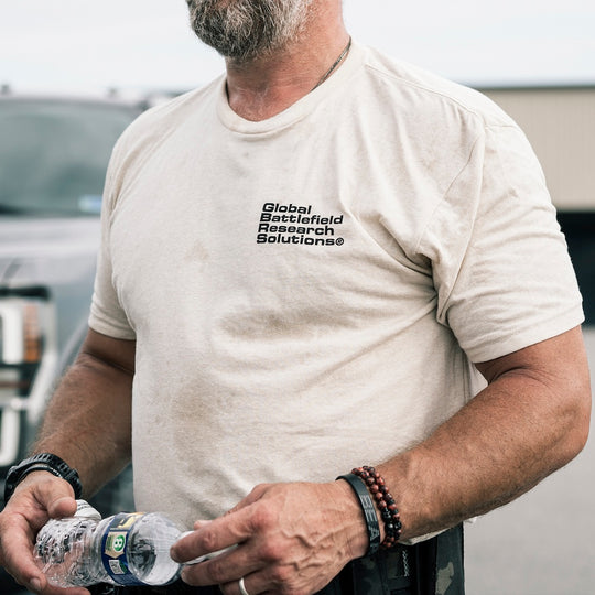 A man wearing the cream GBRS Group Obscured Short Sleeve Shirt with black printed text 'Global Battlefield Research Solutions' on the left chest, holding a water bottle in an outdoor setting.