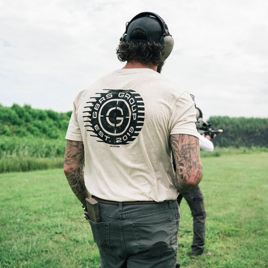 A man standing at a shooting range viewed from behind, wearing a cream t-shirt with a large 'GBRS Group' logo on the back, dark gray pants, and protective earmuffs. Another person is visible in the background of the grassy field.
