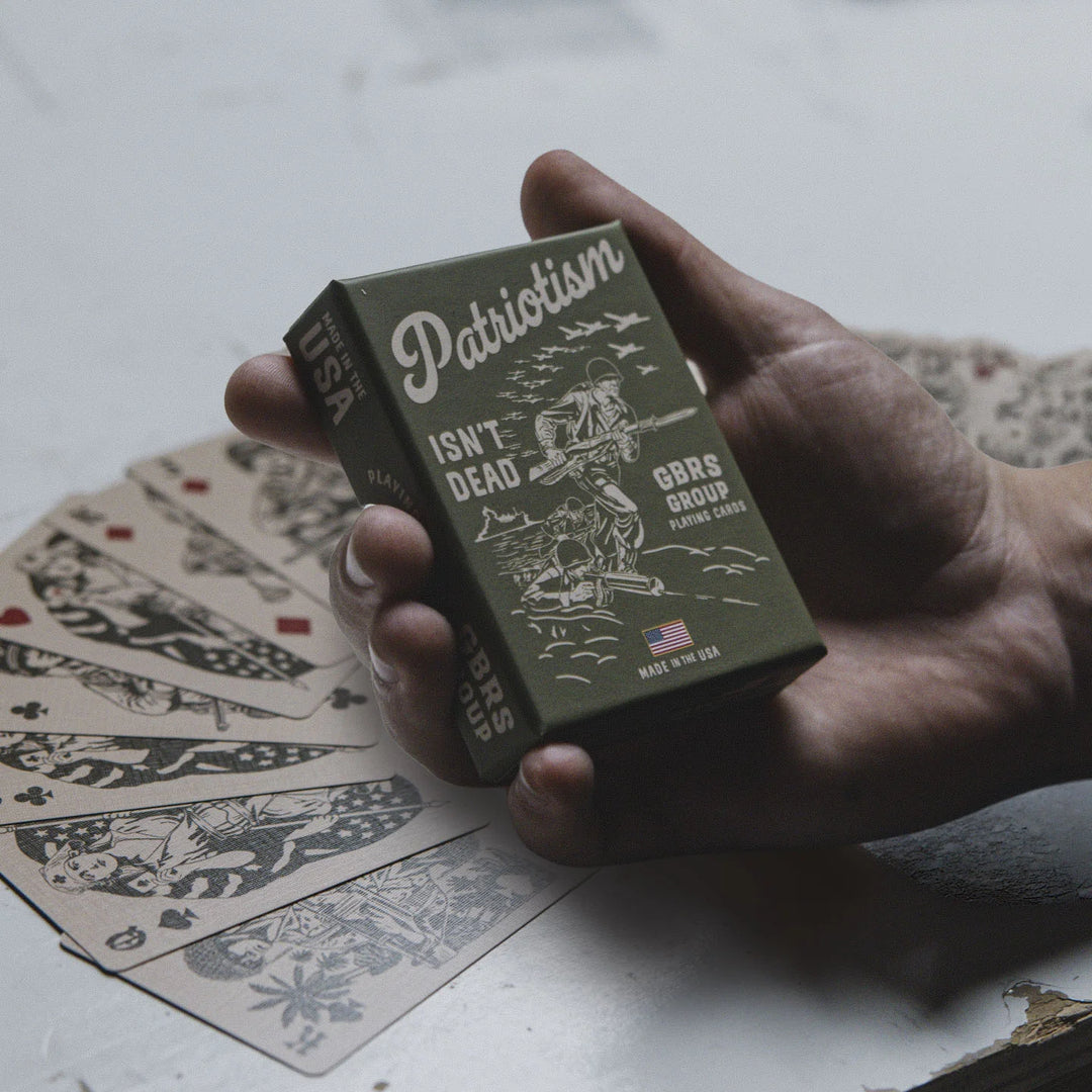 Hand holding a box of playing cards with 'Patriotism Isn't Dead' design on a table.