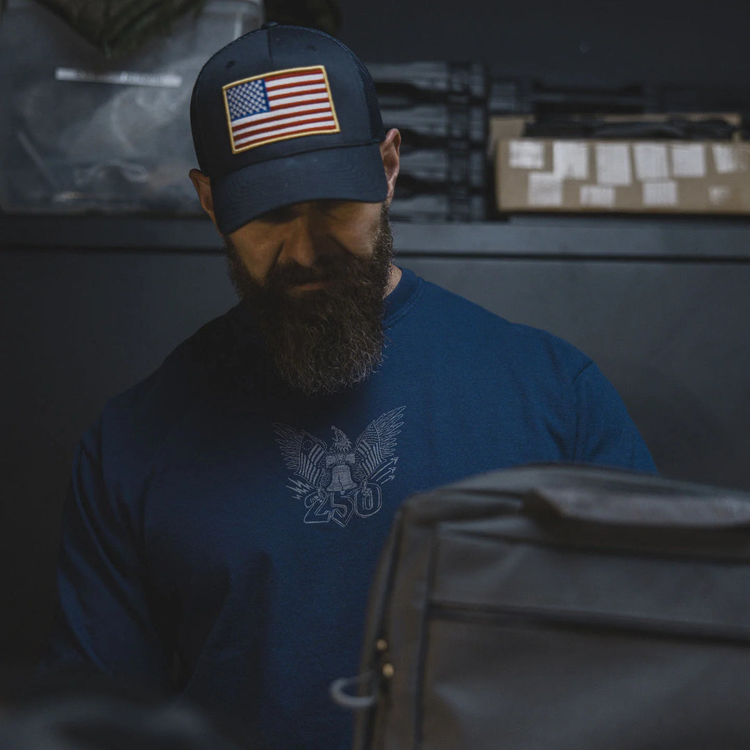 Man wearing a blue cap with an American flag patch and the GBRS Group Semper Fortis Long Sleeve Shirt with an eagle logo, looking at a black bag.