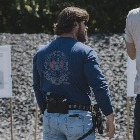 Man wearing the GBRS Group Semper Fortis Long Sleeve Shirt and jeans, holding a firearm, at an outdoor range.